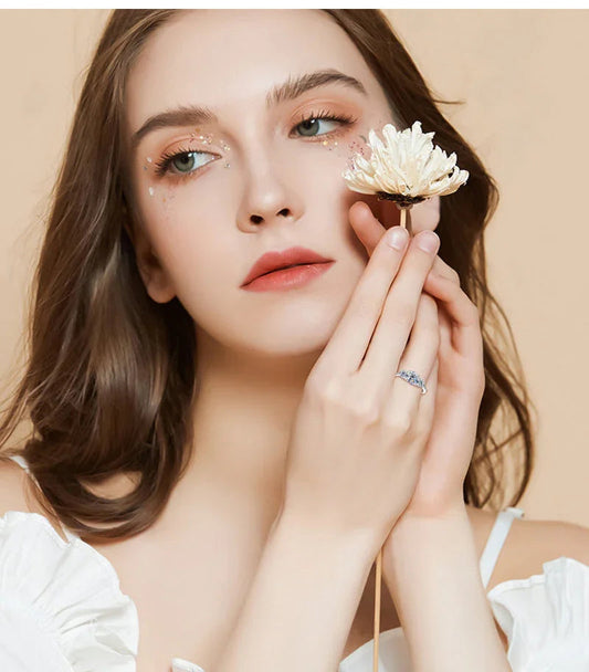 Woman with floral makeup holding ivory flower, wearing a silver ring, neutral background